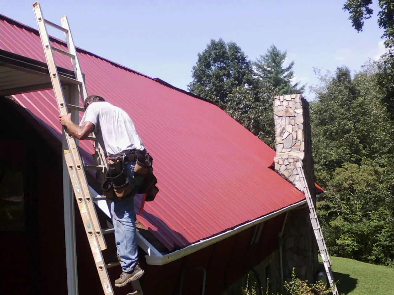 Roofer on a ladder beside a red metal roof and stone chimney — Soffit & Fascia Repair by Roofing Brothers in Hastings On Hudson